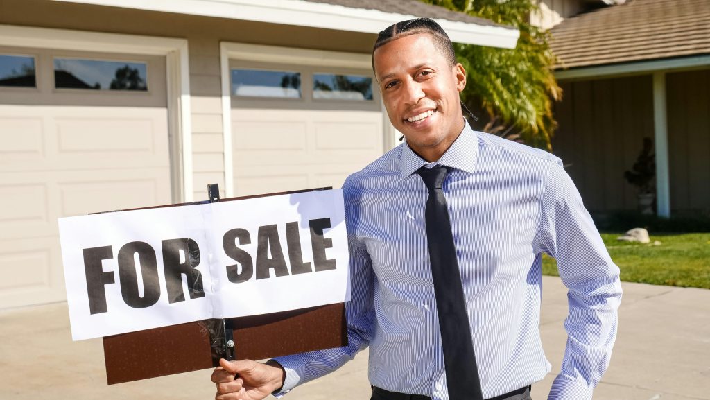 Agent immobilier debout avec un panneau "À vendre" devant une maison par une journée ensoleillée.