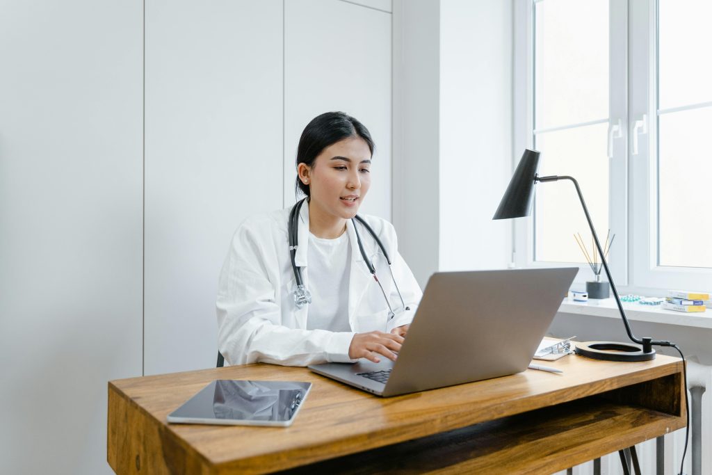 Une femme médecin en blouse blanche utilise un ordinateur portable pour une consultation en ligne depuis son bureau.