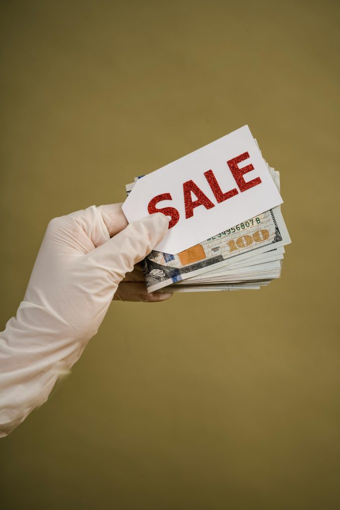 Une main gantée tient une liasse de billets et un panneau de soldes, symbolisant des offres de réduction.