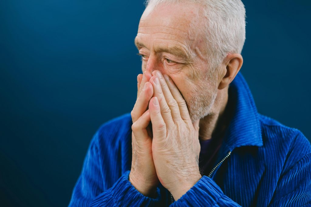 Serieux homme âgé barbu aux cheveux gris en veste bleue couvrant sa bouche avec ses mains tout en regardant ailleurs sur un fond bleu