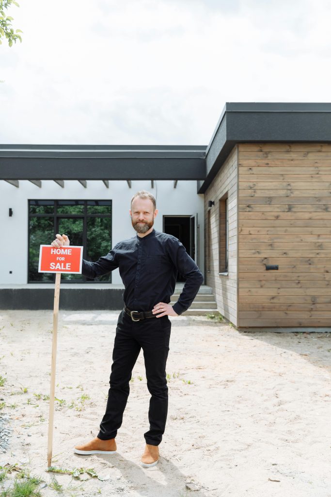 Un agent immobilier se tient à l'extérieur en tenant une pancarte « Maison à vendre » devant une maison moderne.
