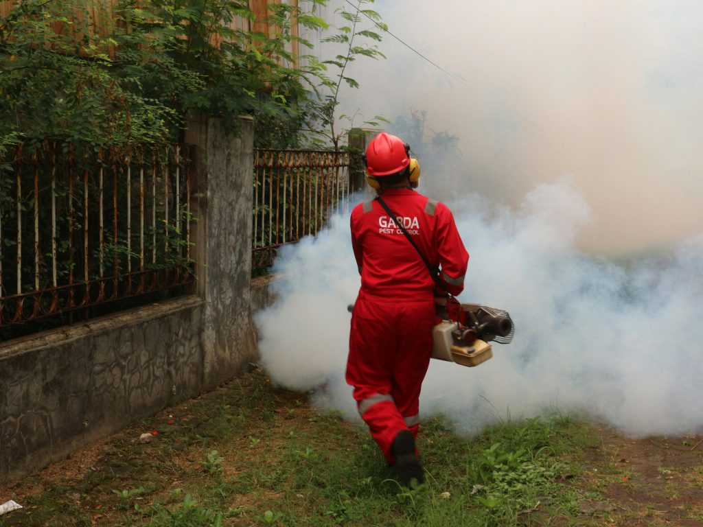 A pest control worker fogging in an outdoor space with smoke to eliminate pests. Un agent de lutte antiparasitaire fumigant un espace extérieur avec de la fumée pour éliminer les parasites.