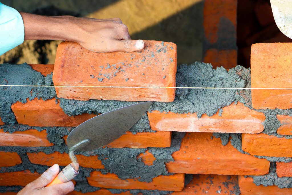 Hands laying bricks with cement in a construction setting, showcasing precise masonry work. Des mains posant des briques avec du ciment dans un environnement de construction, mettant en avant un travail de maçonnerie précis.