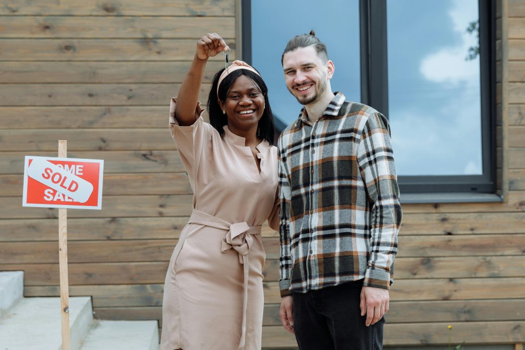 Young couple celebrates buying their first house with keys in hand and a sold sign. Un jeune couple célèbre l'achat de sa première maison avec les clés en main et un panneau vendu.