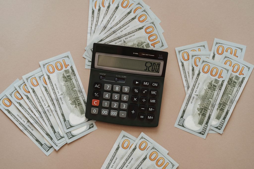 Top view of a calculator showing $5200 surrounded by $100 bills on a tan background. Vue de dessus d'une calculatrice affichant 5200 $ entourée de billets de 100 $ sur un fond beige.