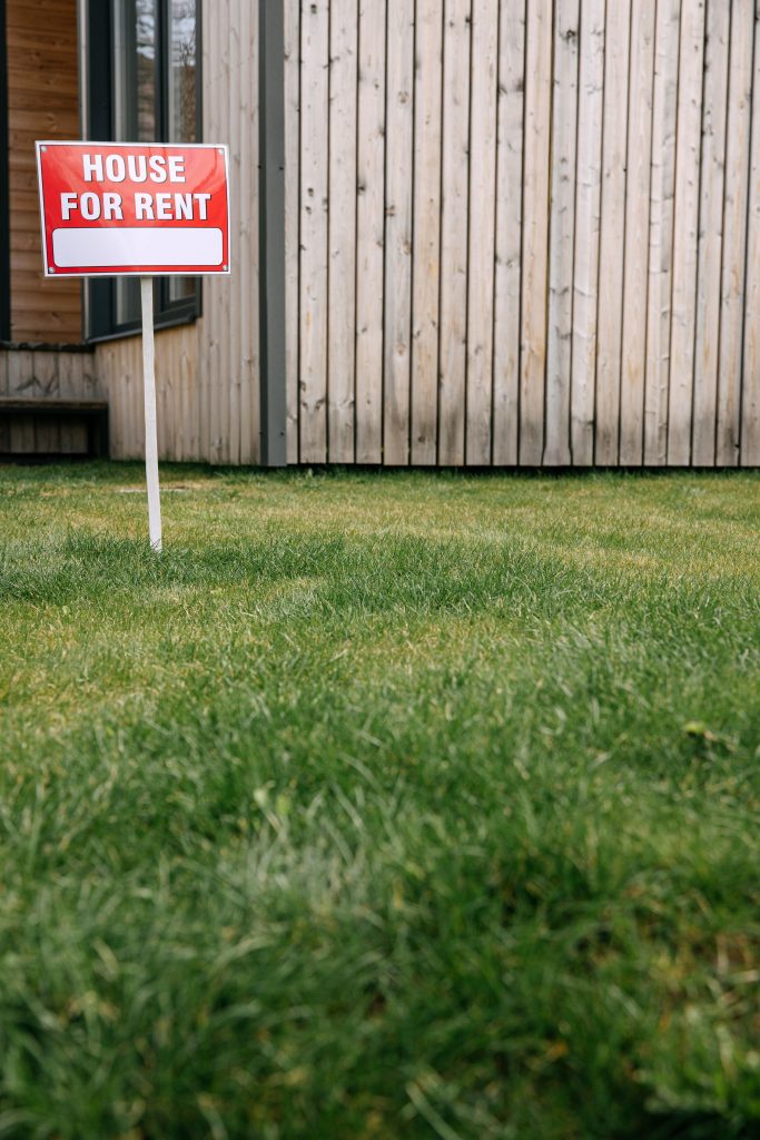 Red 'House for Rent' sign on green lawn with wooden house wall in background. Panneau rouge 'Maison à louer' sur pelouse verte avec mur de maison en bois en arrière-plan.